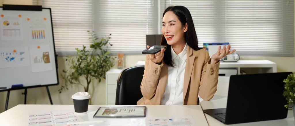 Asian businesswoman in a beige suit talking to voice assistant or chatbot on her smartphone in office, sitting in modern office, using ai technology, smart assistant.